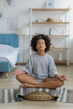 Full body of concentrated African American girl in casual clothing with curly hair meditating on blanket on support with eyes closed in bright cozy room in daytime